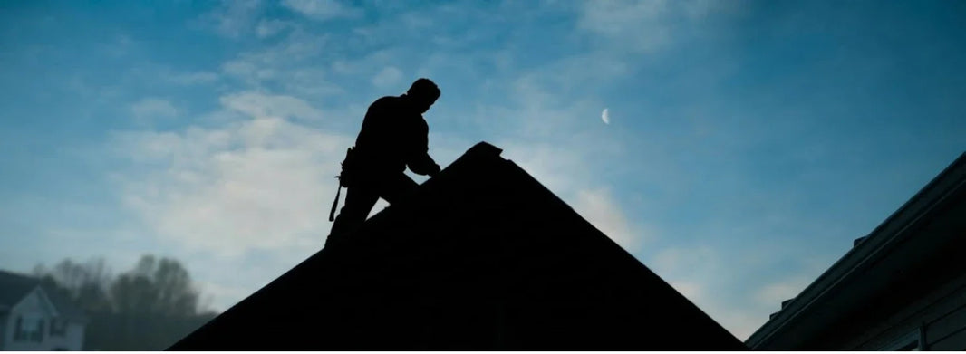 A silhouette of a general contractor on the roof, dark against a dim blue sky.