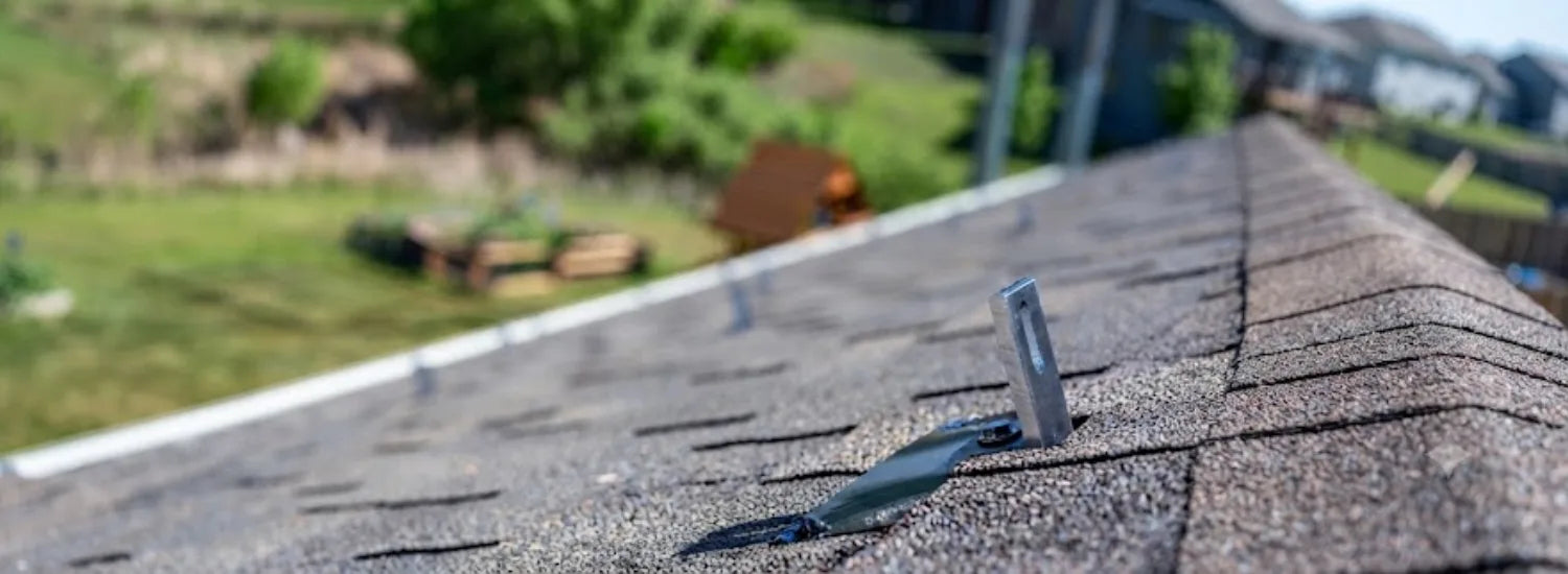 A closeup of an asphalt rooftop, on which a metal roof anchor is attached, and behind which a blurry, green residential neighborhood is visible.