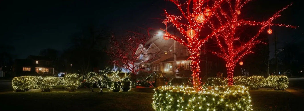 A sumptuous lit, large manor home, with red Christmas lights and hanging lanterns on the trees and bright white Christmas lights on the hedges and home itself.