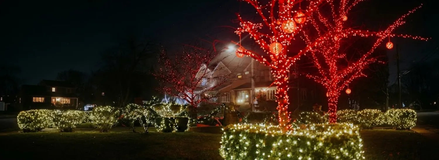 A sumptuous lit, large manor home, with red Christmas lights and hanging lanterns on the trees and bright white Christmas lights on the hedges and home itself.