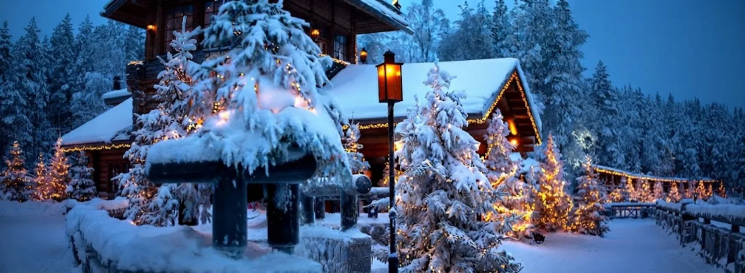 A photo of a snow-covered house in Christmas Village in Finland, which is ringed in warm yellow Christmas lights and obscured by snow-covered firs that surround it. A warmly lit street lantern is visible nearby.