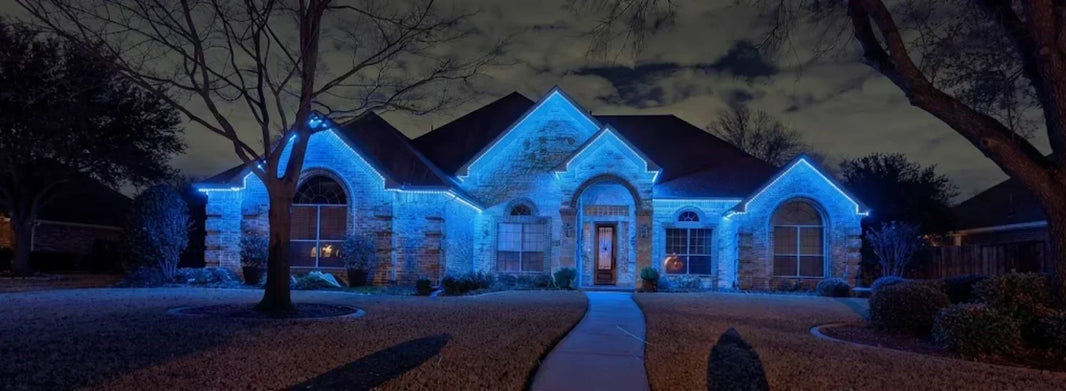 A large, brick-facade house at evening time, thoroughly lit by a single shade of blue Christmas lights along its upper awnings. The surroundings are extremely well lit by the lights, making the walkways visible.